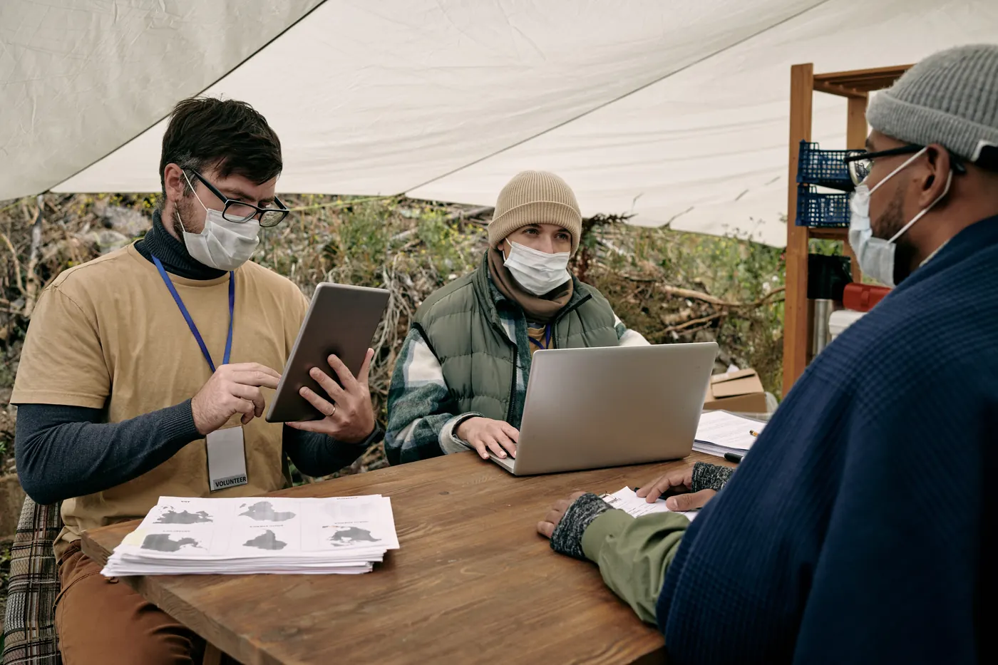 Two volunteers at an outdoor service table, one using a tablet and one a laptop, recording information while they meet with a community member